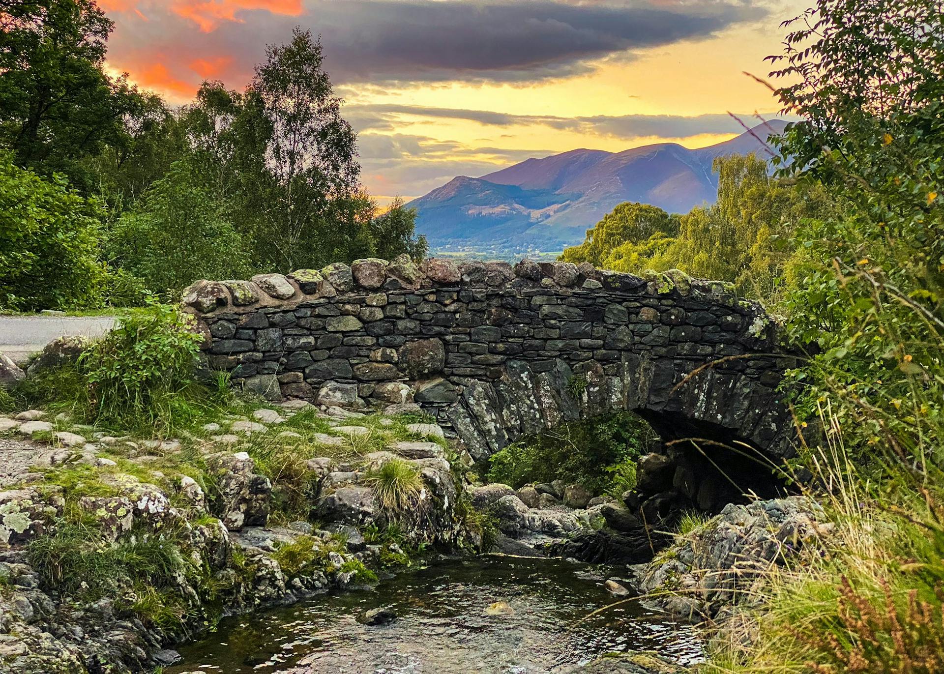 <p>Ashness Bridge is a traditional stone-built bridge on a single-track road in the English Lake District, Cumbria. It is a popular spot for photographers and visitors, as it offers a stunning view of the Borrowdale valley, Derwentwater lake, and Skiddaw mountain. </p><p><br></p><p>The bridge dates back to the 17th or 18th century, and was used by packhorses carrying goods from the hamlet of Watendlath to the town of Keswick. The bridge is owned by the National Trust, and there is a small car park nearby. You can also reach the bridge by taking the Keswick Launch to Ashness Gate, or by taking the bus to Derwentwater Youth Hostel. </p><p><br></p><p>Near the bridge, there is a surprise view point that gives you a panoramic vista of the lake and the town. Ashness Bridge is a beautiful and historic place to visit in the Lake District.</p>